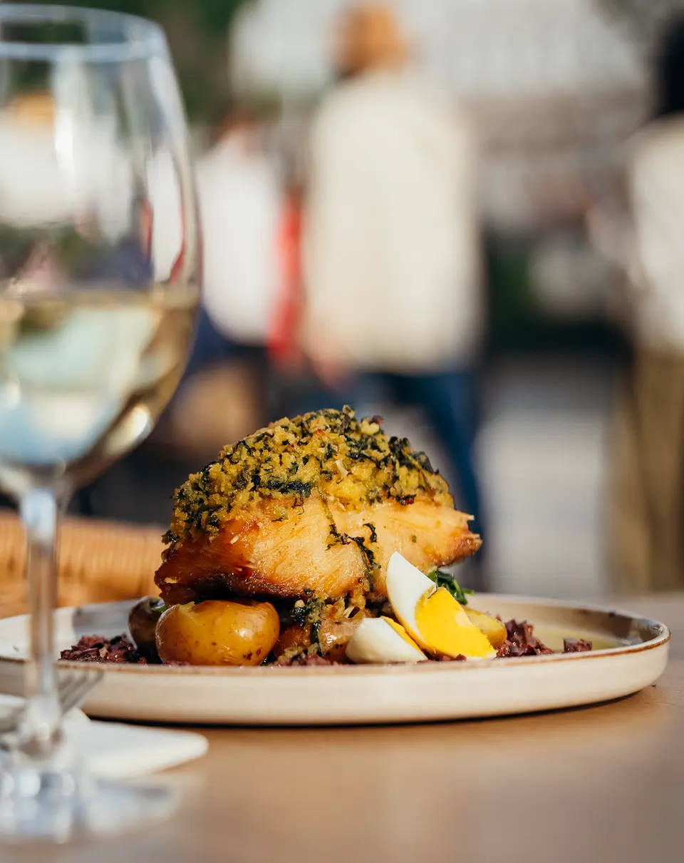 Peixe com crosta de ervas, batatas e um copo de vinho branco, numa esplanada
