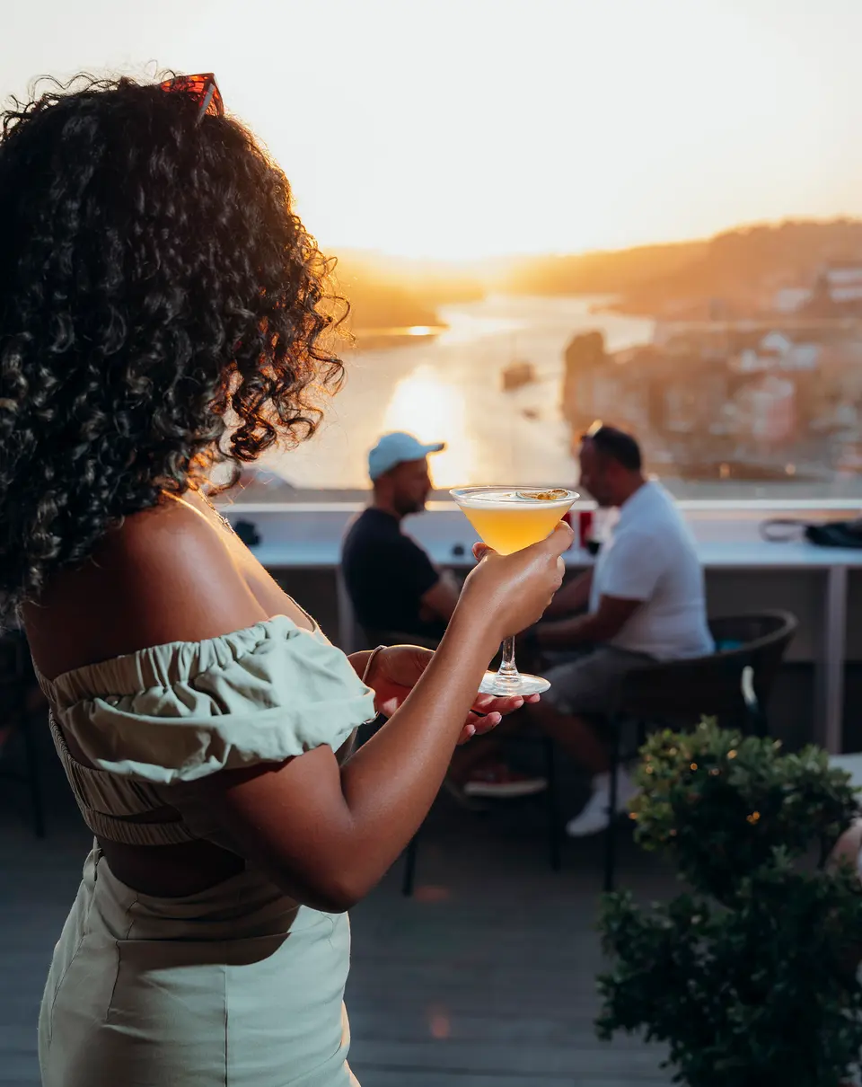 Uma cliente num terraço ao pôr-do-sol sobre o Douro, com um cocktail na mão
