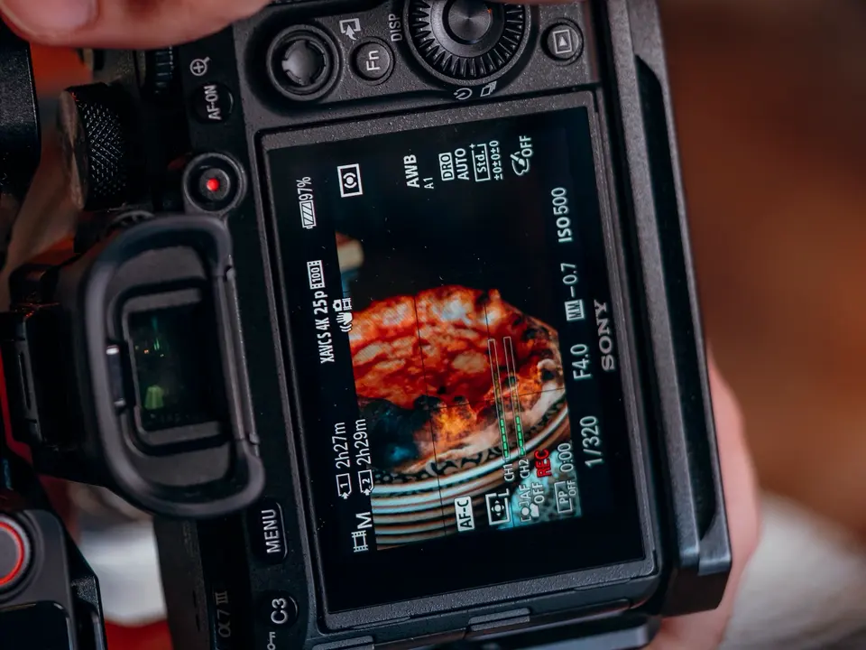O ecrã de uma câmara de filmar a gravar uma pizza, com a mão do fotógrafo por cima