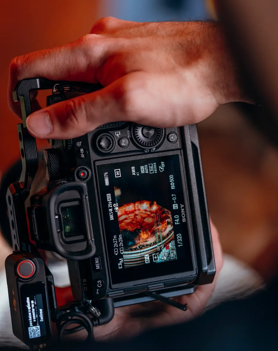 O ecrã de uma câmara de filmar a gravar uma pizza, com a mão do fotógrafo por cima